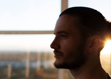 Close-up side portrait of young man on background of sunset and window.