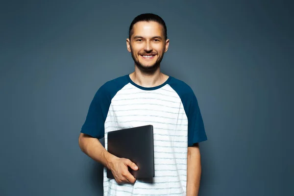 Studio portrait of smiling guy in shirt, holding laptop on blue background.