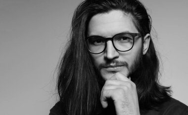 Black and white portrait of young serious man with long hair, wearing eyeglasses, holding hand on chin, grey background.