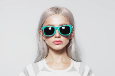 Studio portrait of serious, beautiful young girl with blonde hair in striped shirt on white background, wearing blue sunglasses.