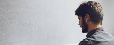 Close-up portrait of young man with wireless earphones on grey textured background with copy space, panoramic banner, back view.