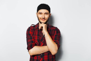 Studio portrait of young smiling guy keeps hand on chin, wearing red shirt and black head band on white background.