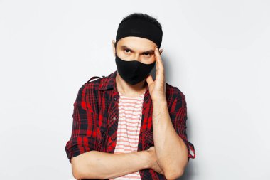 Studio portrait of sad man wearing black protective mask, have headaches, keeps hand on head, on white background.