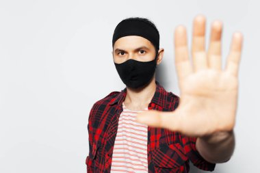 Studio portrait of young man wearing protective mask of black, showing stop gesture by hand on white background.