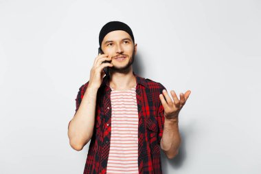 Studio portrait of young cheerful man talking on smartphone on white background, gesturing with hand. Wearing red plaid t-shirt and striped shirt.