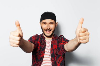 Studio portrait of young joyful handsome man, showing thumbs up, wearing red plaid shirt and black headband.