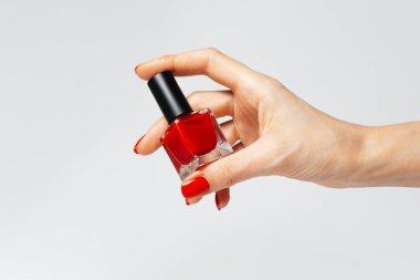 Close-up of female hand with red manicure holding a glitter nail polish of red color on white background.
