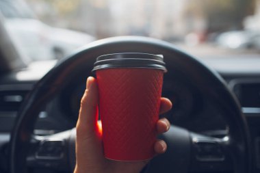 Close-up of male hand holding paper cup with coffee takeaway of red color inside car behind the vehicle wheel.