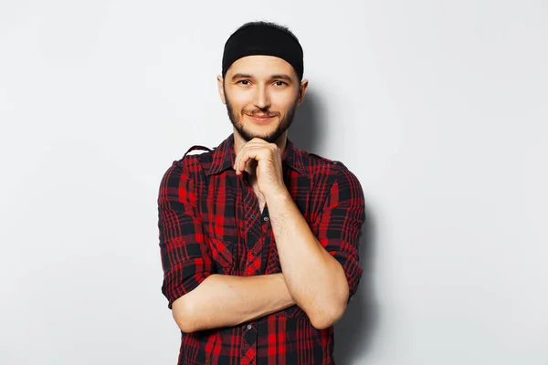 Studio portrait of young smiling guy keeps hand on chin, wearing red shirt and black head band on white background.