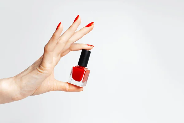 Close-up of female hand with red manicure holding a glitter nail polish of red color on white background.