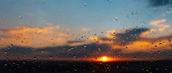 Close-up of raindrops on window on background of beautiful colourful sunset. Natural abstract background. Panoramic banner view.