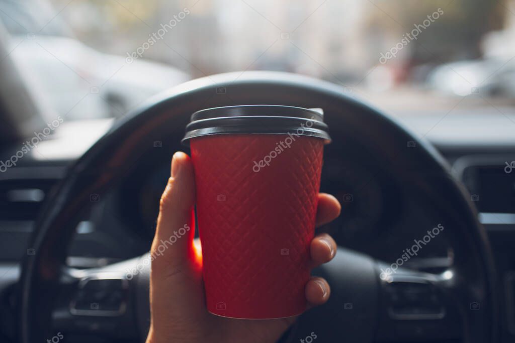Close-up of male hand holding paper cup with coffee takeaway of red color inside car behind the vehicle wheel.