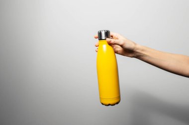 Close-up of female hand holding stainless reusable thermo water bottle of yellow color on grey background.