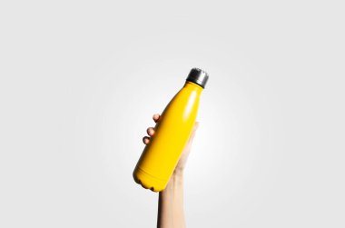 Close-up of female hand holding eco reusable thermo water metal bottle of yellow color on light grey background.