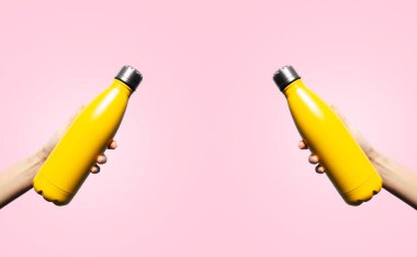 Close-up of female hands holding reusable water bottles of yellow color on pastel pink background.
