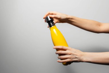 Close-up of female hands holding steel reusable thermo water bottle of yellow color on grey background.