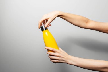 Close-up of female hands holding metal reusable thermo water bottle of yellow color on grey background.
