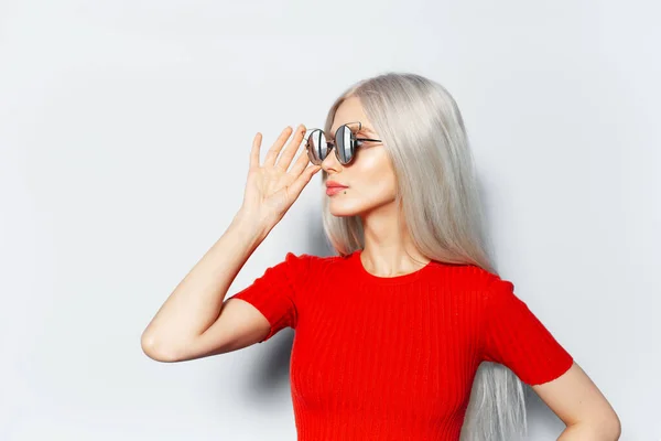 Studio side portrait of young blonde pretty girl wearing sunglasses and red shirt, looking away on white background.