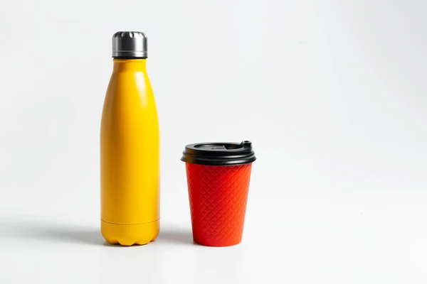 Close-up of metal thermo water bottle of yellow near paper cup for coffee takeaway of red color, on white studio background.