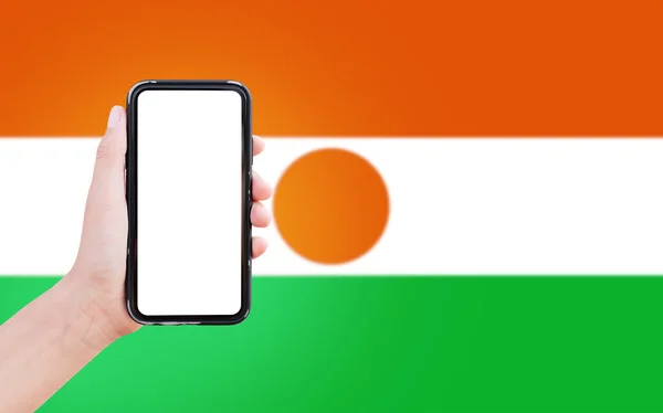 Close-up of male hand holding smartphone with blank on screen, on background of blurred flag of Niger.