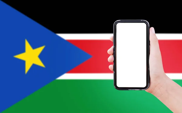 Close-up of male hand holding smartphone with blank on screen, on background of blurred flag of South Sudan.