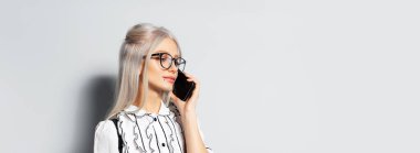 Studio portrait of pretty smiling girl with blonde hair, talking on smartphone, wearing eyeglasses on white background, panoramic banner view with copy space.