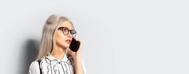 Studio portrait of pretty girl with blonde hair, talking on smartphone, wearing eyeglasses on white background, panoramic banner view with copy space.