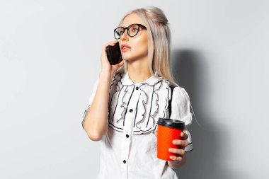 Studio portrait of young cute girl with blonde hair using smartphone, holding paper cup of coffee takeaway, wearing eyeglasses on white background.
