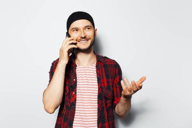 Studio portrait of young cheerful man talking on smartphone on white background, gesturing with hand. Wearing red plaid t-shirt and striped shirt.