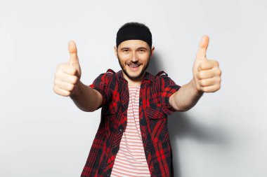 Studio portrait of young joyful handsome man, showing thumbs up, wearing red plaid shirt and black headband on white background.