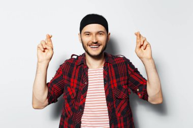 Studio portrait of young handsome happy man, with crossed fingers, wearing red plaid shirt,  on white background.