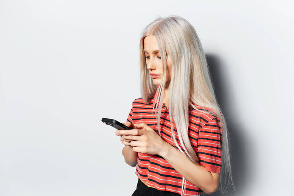 Студийный портрет молодой красивой девушки с светлыми волосами, смотрящей в смартфон, в красной полосатой рубашке на белом фоне.