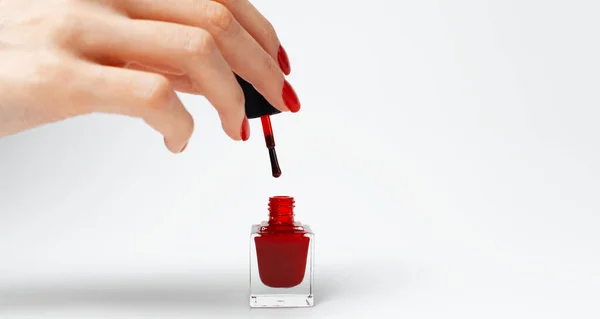 Close-up of female hand with red manicure holding a glitter nail polish of red color on white background.