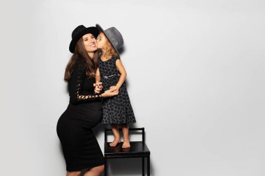 Studio portrait of pregnant smiling woman and child girl kisses her mother on cheek, standing on chair, wearing hats on white background.