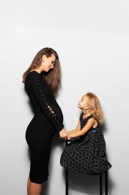 Studio portrait of young mother and her daughter looking each ever, holding hands, wearing black dresses on white background. Pregnant woman.