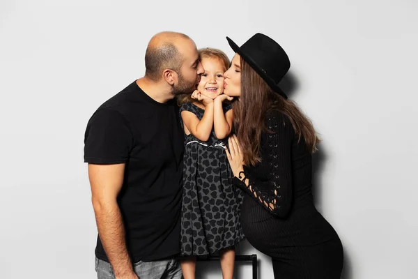 Studio portrait of happy young family. Young pregnant woman in black and a man kisses on cheeks they daughter on white background.