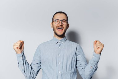 Mavi gömlek ve gözlük takmış, elleri beyaz arka planda zafer jesti yapan mutlu genç adamın stüdyo portresi..