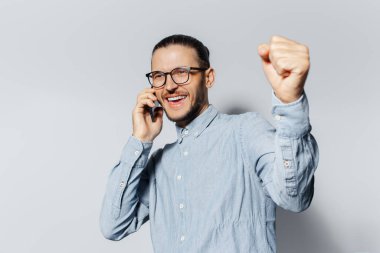 Öyle mi? Akıllı telefondan konuşan genç mutluluğun stüdyo portresi, beyaz arka planda kazanan bir jest yapıyor. Gözlüklü ve mavi gömlekli..