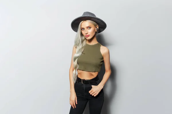 Studio portrait of young beautiful blonde girl with wireless earphones in ear, wearing grey hat on white background.