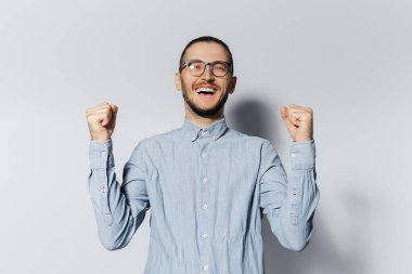 Mavi gömlek ve gözlük takmış, elleri beyaz arka planda zafer jesti yapan mutlu genç adamın stüdyo portresi..