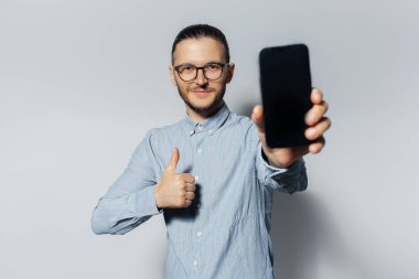 Akıllı telefon ve açık gri arka planda baş parmağını gösteren kendine güvenen genç bir adamın stüdyo portresi.