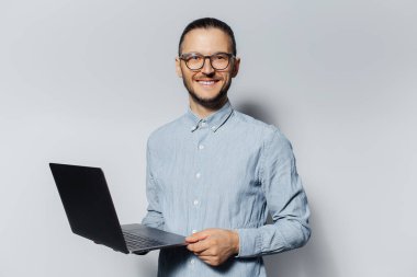 Stüdyoda elinde dizüstü bilgisayarla beyaz arka planda mavi gömlek ve gözlük takan genç bir adamın portresi..