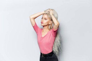 Stüdyo portresi, genç sarışın çekici kız, elleri başının üstünde, beyaz arka planda pastel pembe gömlek giyiyor..