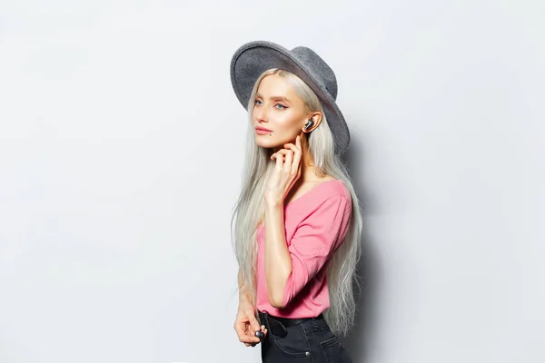 照片上年轻漂亮的金发姑娘 耳朵里戴着无线耳塞 头戴灰色帽子 身穿粉色衬衫 背景为白色 — 图库照片