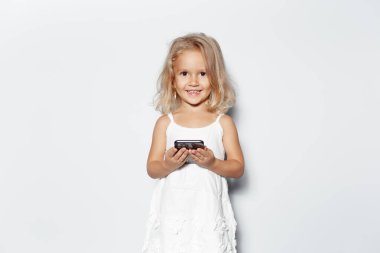 Beyaz arka planda elinde akıllı telefonla gülümseyen sarı saçlı çocuk portresi..