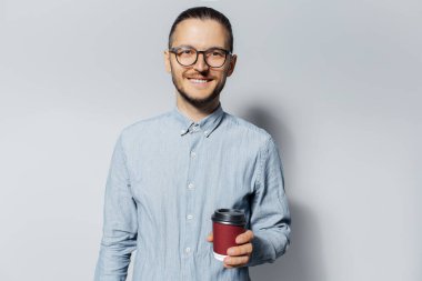 Elinde kahve fincanı olarak beyaz arka planda kırmızı kağıt bardak tutan genç bir adamın stüdyo portresi. Mavi gömlek ve gözlük takıyor..