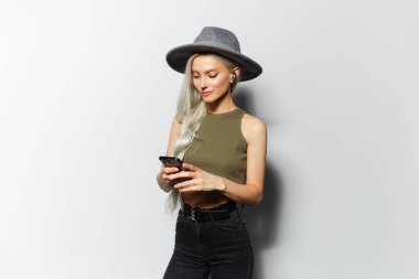 Akıllı telefon ve kablosuz kulaklık kullanan, beyaz arka planda gri şapka takan güzel sarışın kızın stüdyo portresi..