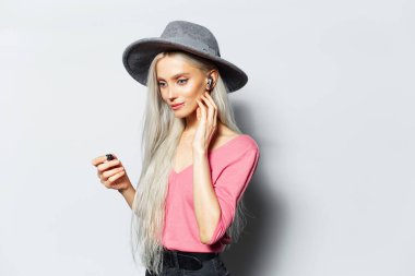 Kulaklıkla müzik dinleyen, beyaz arka planda gri şapka ve pembe gömlek giyen sarışın güzel bir kızın stüdyo portresi..