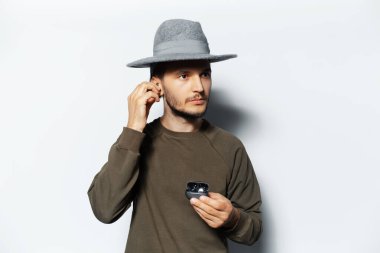 Kulak kablosuz kulaklık takan, beyaz arka planda şarj çantasını tutan, süveter giyen ve gri şapkalı genç bir adamın stüdyo portresi..