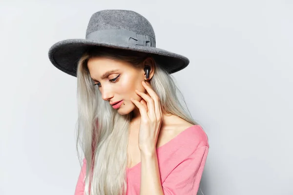 年轻金发可爱女孩通过耳塞听音乐的工作室肖像 背景为白色 头戴灰色帽子 身穿粉色衬衫 — 图库照片
